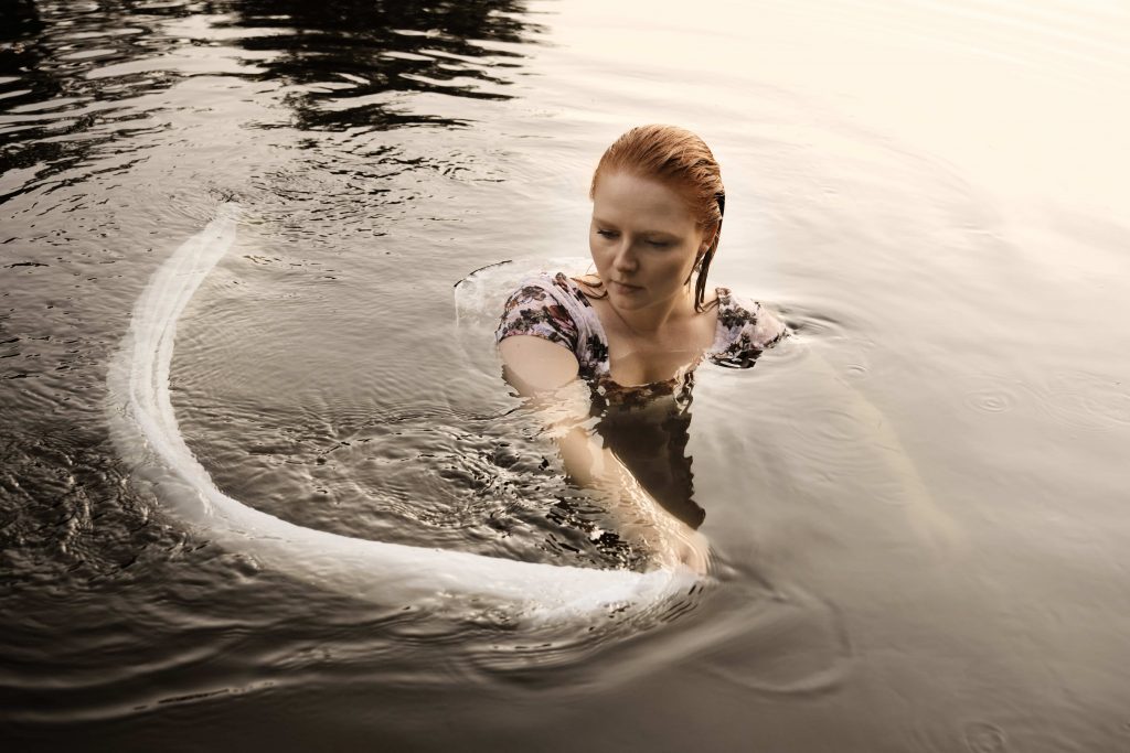 Ivory Stone during sunset, submerged up to the shoulders in water of a river, moving a long piece of silken cloth through the water in a spiral motion