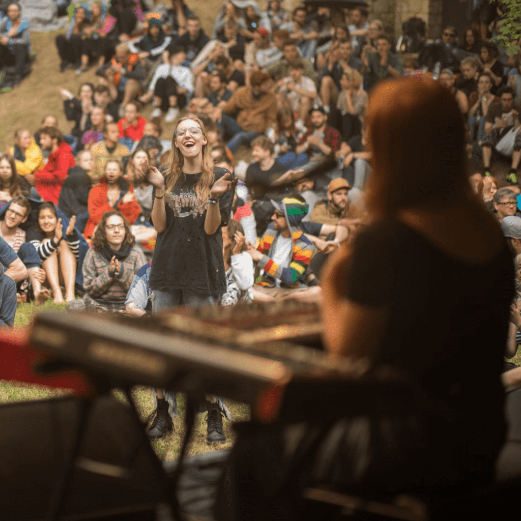 Photograph taken at an open-air concert of Ivory Stone, view from behind the stage onto the artist sitting at the piano. The audience in the background sits and claps, one audience member in the front is standing, clapping and cheering enthusiastically.